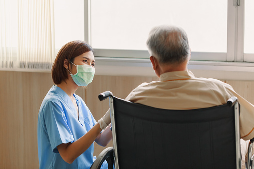 Young Asian woman nurse explaining information to elderly man patient in wheelchair with friendly smiley face in the hospital. Young Assistance with old people in the elderly care place.