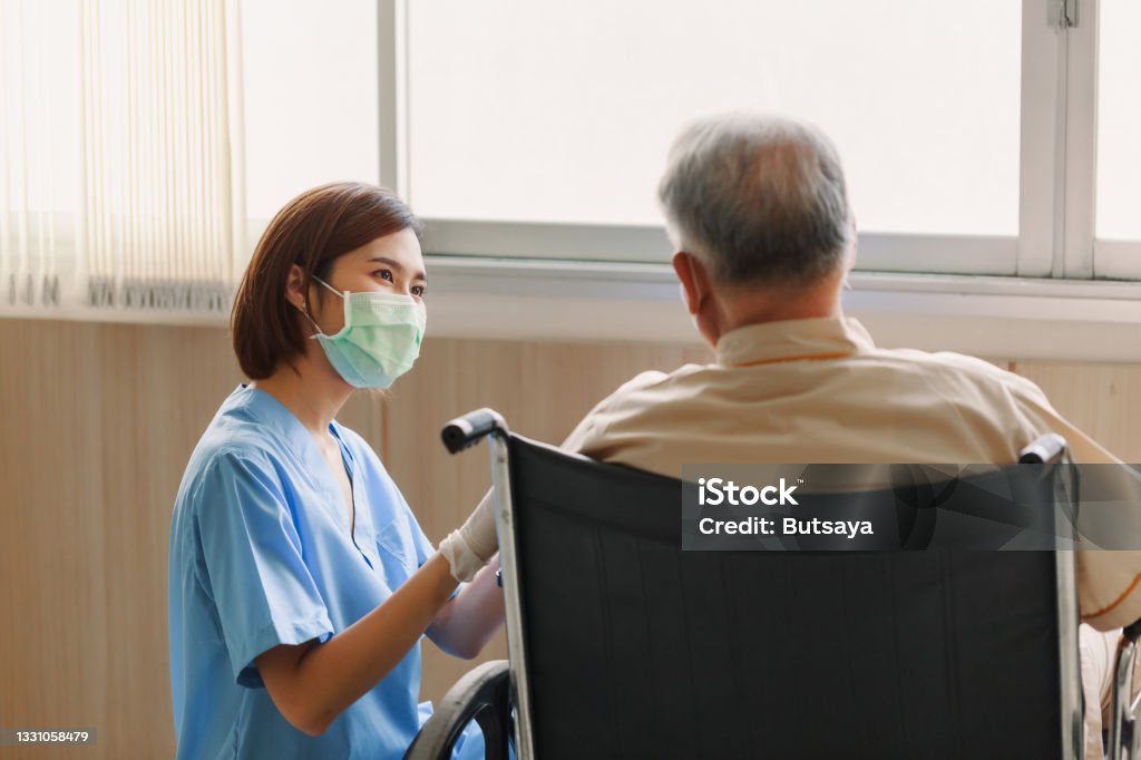 Young Asian woman nurse explaining information to elderly man patient in wheelchair with friendly smiley face in the hospital. Young Assistance with old people in the elderly care place Young Asian woman nurse explaining information to elderly man patient in wheelchair with friendly smiley face in the hospital. Young Assistance with old people in the elderly care place. Protective Face Mask Stock Photo