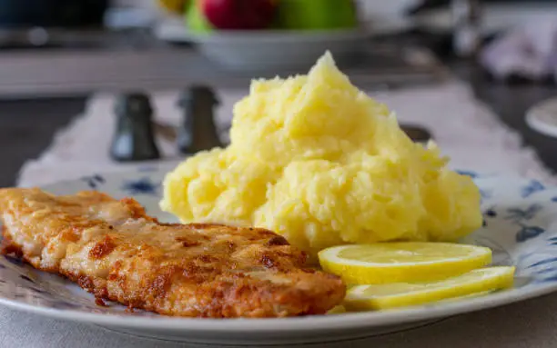 Traditional fresh cooked fried fish with delicious homemade mashed potatoes served on a plate with sliced lemon. Closeup and front view with blurred table background