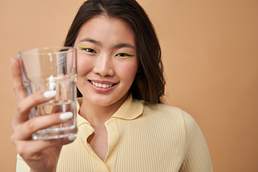 Do you want. Portrait view of the asian woman stretching transparent glass of water to the camera while purposing beverage for somebody. Stock photo