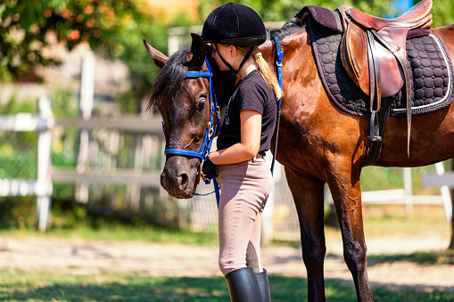 Beautiful girl in riding gear holds her horse by the reins. She stroking him after class at riding school.