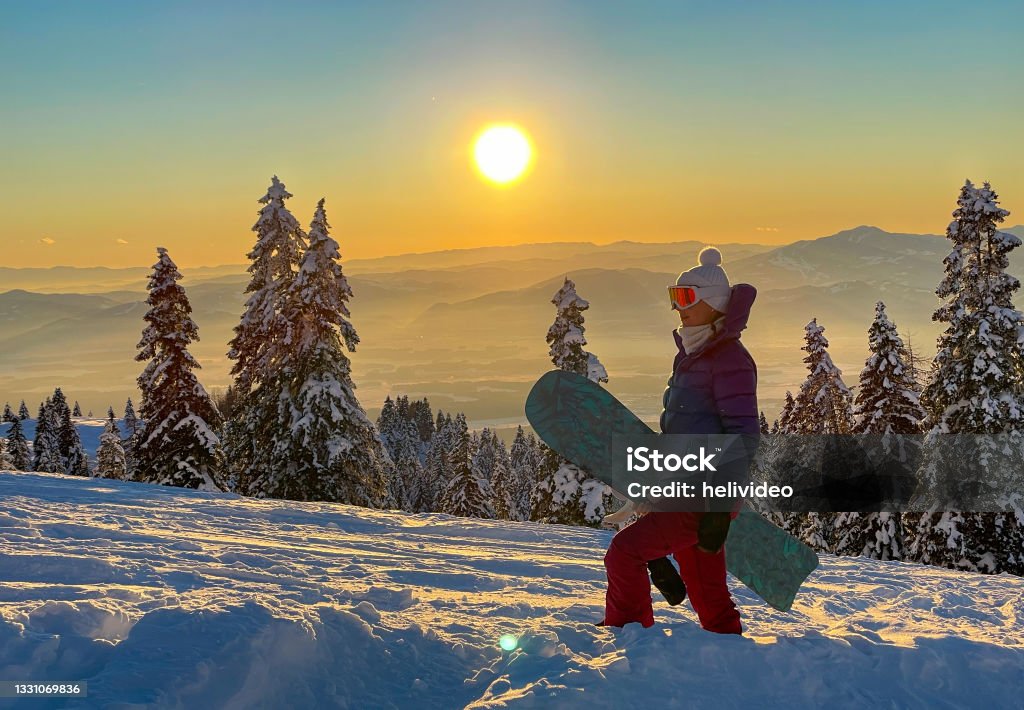 CLOSE UP: Young female snowboarder hikes up ungroomed snowy slope at sunset. CLOSE UP, LENS FLARE: Young female snowboarder hikes up ungroomed snowy slope at sunset. Golden winter morning sun rises above the idyllic snowy mountain and woman on snowboarding trip hiking up hill. Beauty Stock Photo