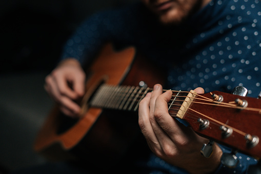 Close-up of talented guitarist singer male playing acoustic guitar sitting on armchair in dark living room, selective focus. Creative musician enjoying leisure activity in apartment.