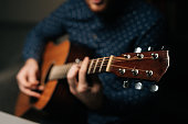 Close-up of unrecognizable guitarist singer male playing acoustic guitar sitting on armchair in dark living room, selective focus.