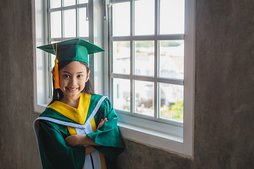 Portrait of cute Asian little girl wearing graduation hat and gowns standing in classroom at school, Elementary School and students concept
