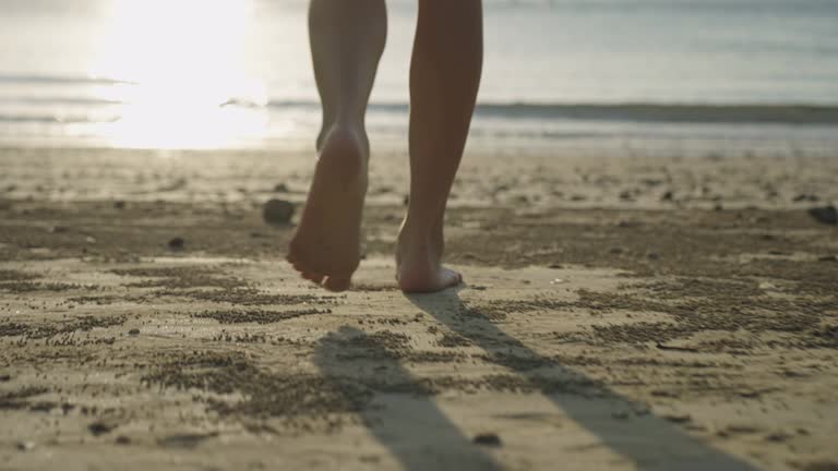 Female legs and feet walking down toward sea water waves on sandy beach, during sunset, relaxing walk escape from busy city, island paradise retreat, low angle shot from behind in slow motion