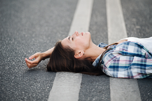 Serene woman lying on an asphalt road, relaxing, looking up at the sky. Side view. She's wearing checkered shirt over regular white.