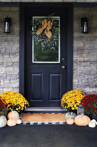 Front porch decorated for Thanksgiving Day with homemade wreath hanging on door. Heirloom gourds,  white, green and orange pumpkins, and colorful mums give an inviting atmosphere.