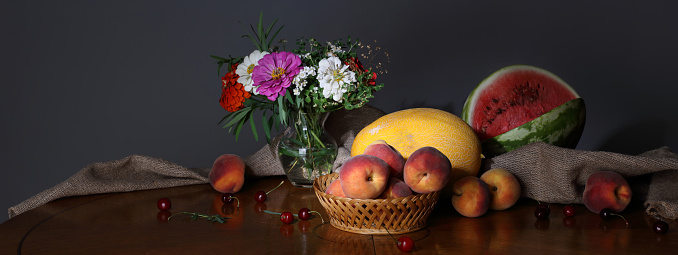 Large-format panorama with a bouquet of summer flowers, peaches, watermelon and melon on a dark background