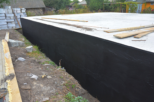 Basement and foundation construction and waterproofing by applying asphalt, bitumen or rubber based tar and digging a drainage trench. Exterior foundation walls sprayed with tar.
