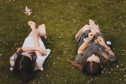 Top photo of two little girls in graceful dresses lying on the grass with delicate flowers in their hands. Friends having fun together outdoors. Bouquet of flowers in the hands of a child.