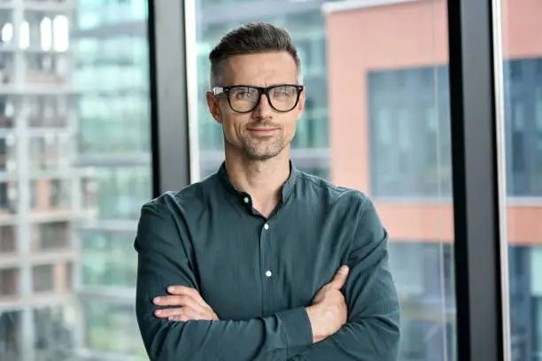 Smiling confident mature businessman looking at camera standing in office. Elegant stylish corporate leader successful ceo executive manager wearing glasses posing for headshot business portrait.