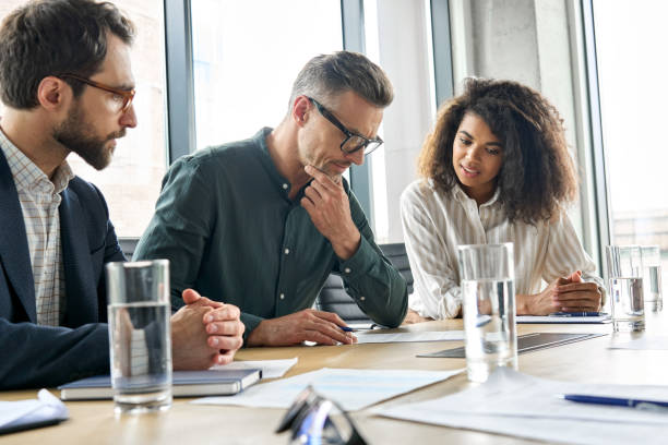 Focused mature businessman reading contract considering risks with lawyers. Focused doubtful mature businessman reading contract document thinking considering risks with professional lawyers legal experts executive team analyzing financial report sitting at office table. decisions stock pictures, royalty-free photos & images