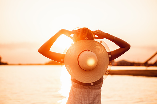 Photo a rear view of a young woman with a hat. A beautiful happy blonde in a lace summer dress and a straw hat decorated with a ribbon stands in the water on the beach against the sunset background.