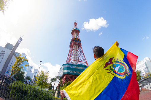 Young latin ecuadorian backpacker tourist fan,  traveling, and celebrating an award on an International sport event with his ecuadorian flag in front of Sapporo Tower at Sapporo Downtown, Hokkaido, Japan. He's wearing a black t-shirt, a black short pants and black boots.\n\nGames fan celebrating concept.\nLatin Male Portrait concept.\nTravel around Japan.\nTourist Concept.