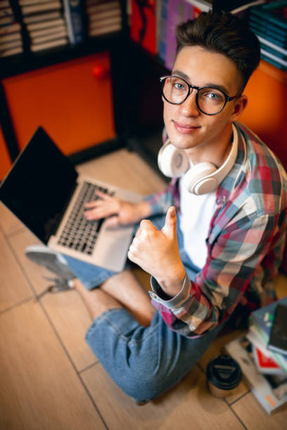 Studying, doing homework. One young caucasian man, student in glasses sitting at floor with laptop at home or library background. Education, studying and student life concept.