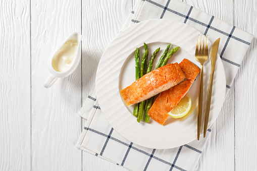 roasted salmon fillets with grilled asparagus and lemon wedges on a white plate, on a white wooden table with hollandaise sauce, horizontal view from above, flat lay