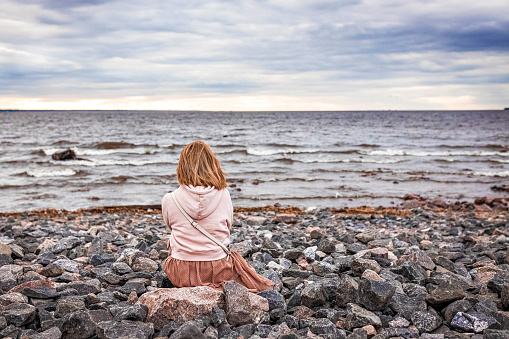 A young woman sits on the rocks on the shore and looks at the evening sea.