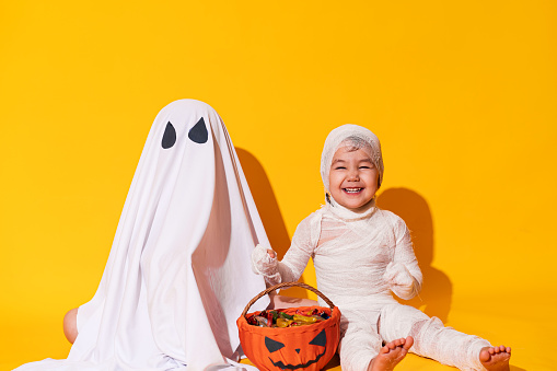 Children in halloween costumes sitting in front of basket of sweets on yellow background.