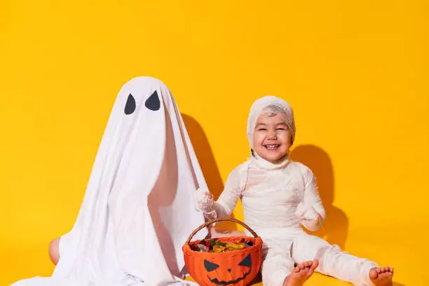 Photo of Children in halloween costumes sitting in front of basket of sweets on yellow background