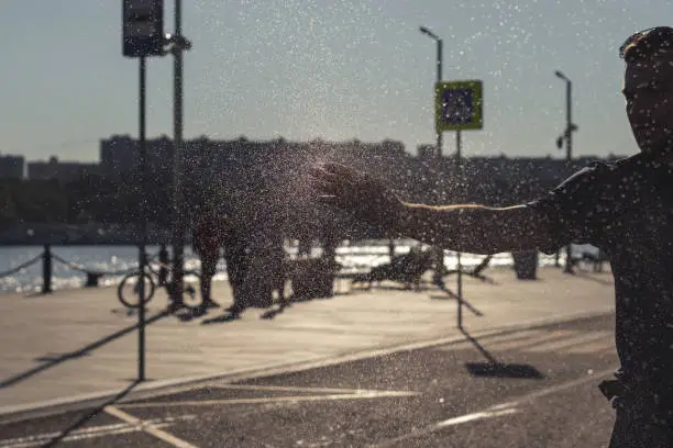 Photo of Water drops in motion. Young man walking on the street and catches water drops with his hand. Water drops in motion. Soft focus on man on background. Summer enjoyment.