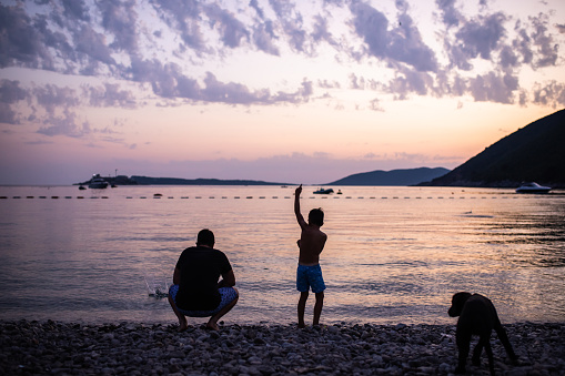 A young father and his small son are stone skipping at the beach at sunset with their puppy