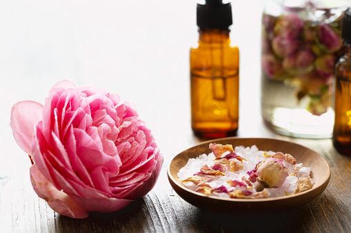 A pink rose covered with drops of watrand sea salt scented with rose aroma, in wooden bowls, mixed with dried rose petals and rose buds, on wooden background, in the background a glass bottle filled with massage oil and dried rose buds  and brown bottles with essential oil