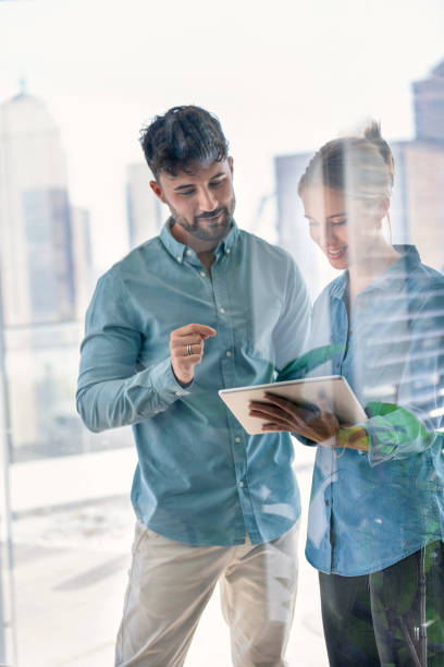 Businessman and businesswoman looking at a digital tablet. Businessman and businesswoman looking at a digital tablet. They are wearing matching casual clothes that could be a uniform and are happy and smiling. Looking through window with reflections two men same clothes stock pictures, royalty-free photos & images