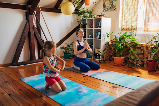 Active mother, teaching her daughter how to meditate in lotus yoga position, while both doing some mother-daughter activity and bonding over yoga