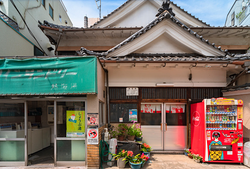 tokyo, japan - july 15 2021: Traditional Japanese bathhouse Atamiyu in Kagurazaka with a beverages vending machine and flowers in front and a coin laundry store with prevention posters beside.