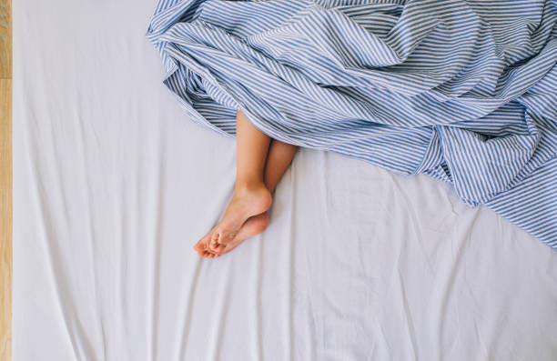 This is a close - up image showing the feet of a person lying under white bedding. The feet are partially covered by the soft white sheets, creating a sense of relaxation and comfort, suggesting a moment of rest or sleep in a cozy environment