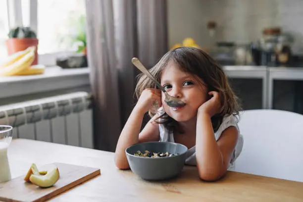Happy young girl having fun while eating cereals with fruit for breakfast in a kitchen in the morning