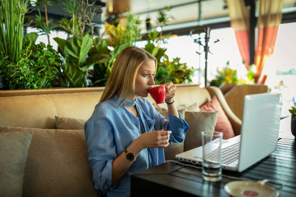 A Young Beautiful Woman is Using her Laptop while Sitting in the Modern Restaurant and Refreshing with a hot Drink.