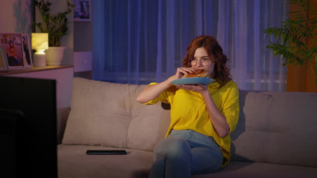 improper diet, beautiful woman with glasses watches TV while eating burger while sitting on couch in room at night, stay at home