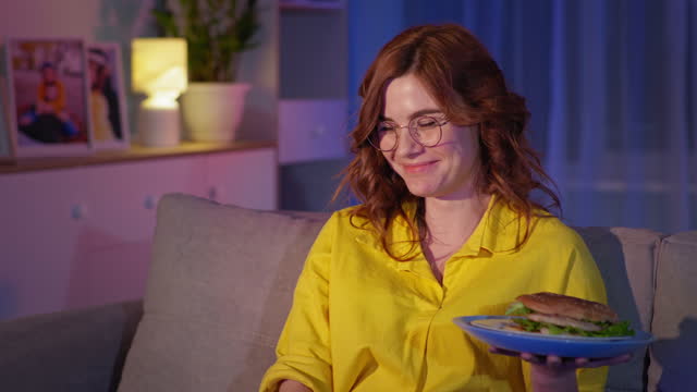 lifestyle, young woman with glasses resting at home in quarantine and eating burger while watching an entertainment show or comedy sitting on couch, stay at home