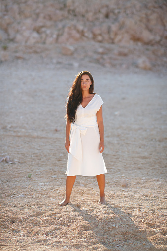 Beautiful brunette in white summer dress standing alone on gravel in stone desert and looking at camera, time alone in nature