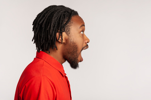 Side view portrait of bearded man with dreadlocks wearing red casual style T-shirt, standing with open mouth, being impressed of shocking news. Indoor studio shot isolated on gray background.