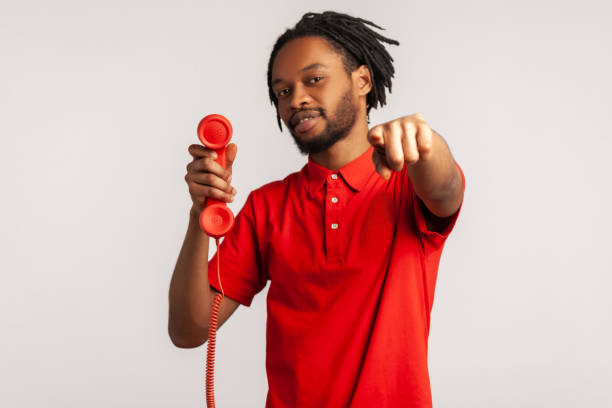 Handsome guy man with dreadlocks wearing red T-shirt, pointing finger to camera, holding handset of red vintage landline phone, answering calls.