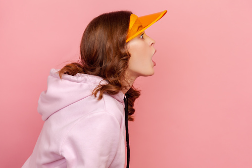 Profile of surprised curly haired teenage girl in hoodie and yellow sun visor cap looking with shocked expression to side. Indoor studio shot isolated on pink background