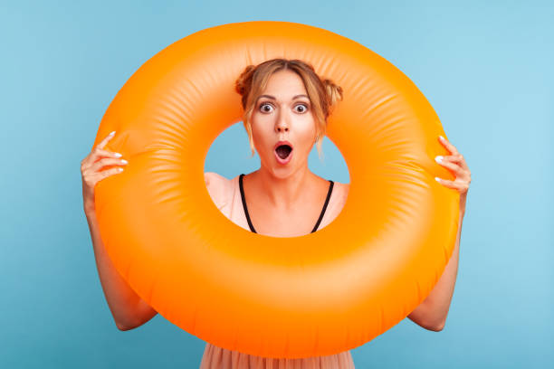 Shocked female with blonde hair and two hair buns holding rubber ring over her neck and looking at camera with surprised expression and opened mouth. Shocked female with blonde hair and two hair buns holding rubber ring over her neck and looking at camera with surprised expression and opened mouth. Indoor studio shot isolated on blue background. condom photos stock pictures, royalty-free photos & images