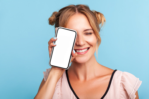 Portrait of joyful blonde female with two buns hairstyle in summer dress, smiling and covering half face with cell phone, looks playful and carefree. Indoor studio shot isolated on blue background.