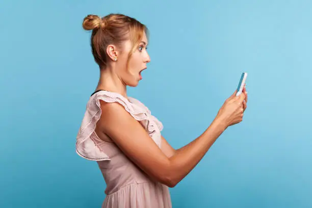 Photo of Side view of surprised girl in stylish dress, expressing shock while using cell phone, online mobile application, searching web, astonishing news.