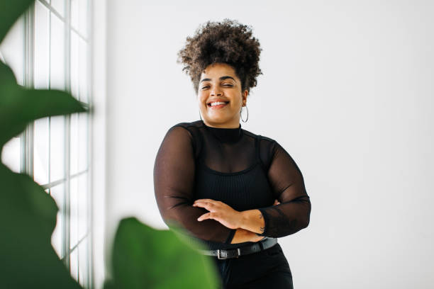 Lady in fashionable outfit standing near window Beautiful woman standing in living room with her arms crossed looking at camera. Female in black outfit standing near window. plus size stock pictures, royalty-free photos & images