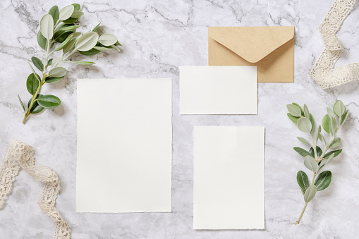 Wedding stationery set with envelope laying on a marble table decorated with eucalyptus branches and ribbons. Mock-up scene with blank paper greeting cards. Feminine flat lay