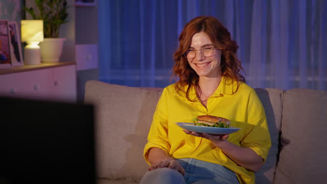 junk food at night, hungry adult girl with glasses watches funny show and eats hamburger during an evening rest at home in quarantine