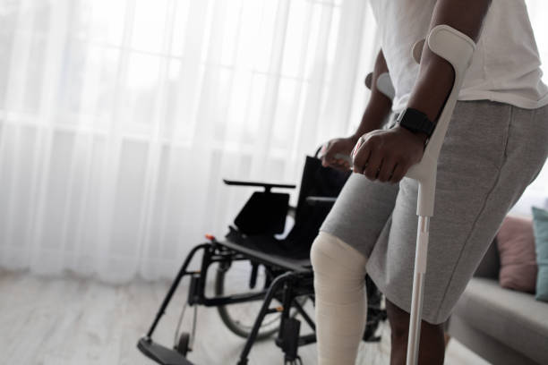 Trauma, post-incident fracture of bone, exercise and recovery at home Trauma, post-incident fracture of bone, exercise and recovery at home. Tense millennial african american man with broken leg in cast walks with crutches to wheelchair in living room interior, cropped black crash stock pictures, royalty-free photos & images