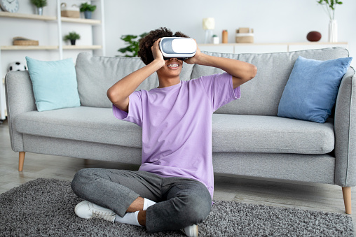 Full length of black teen guy wearing VR headset and enjoying cyberspace exploration at home. African American adolescent absorbed in virtual reality. Modern technologies in everyday life concept