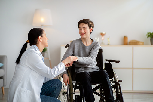 Disabled people healthcare support concept. Doctor holding hands of handicapped teen boy in wheelchair during medical visit. Happy impaired adolescent and young nurse at home