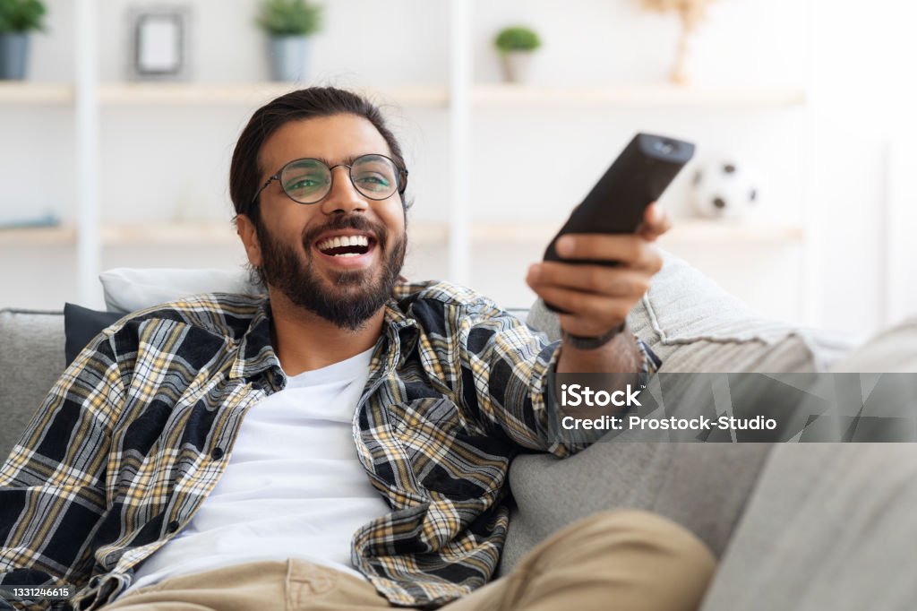 Relaxed arab guy wathcing movie on TV Relaxed arab guy wathcing movie on TV at home, sitting on couch alone, having happy face expression. Happy young man with TV remote watching comedy and laughing, copy space, closeup Remote Control Stock Photo