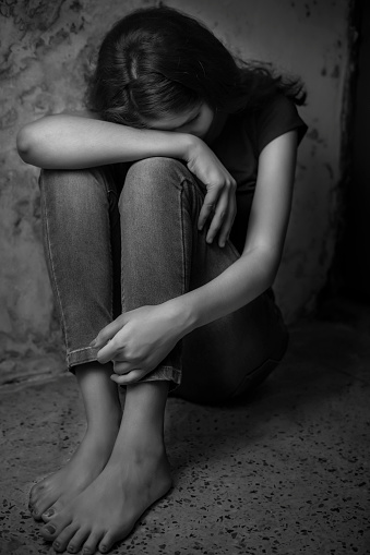 Black and white portrait of a sad young woman sitting by hiding herself in corner of a dark room near the dirty wall.